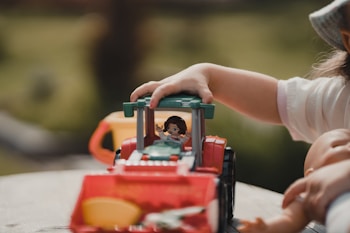 A child is playing with a toy tractor on a wooden surface. Their hand is positioned on the toy, which is brightly colored in red and green. There's a small figure inside the driver's seat of the tractor. The background is out of focus, suggesting an outdoor setting with greenery.