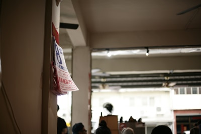 Posters with information in a foreign language are attached to a column inside an open space with visible industrial elements. Various indistinct figures and a busy street can be seen through a large opening.