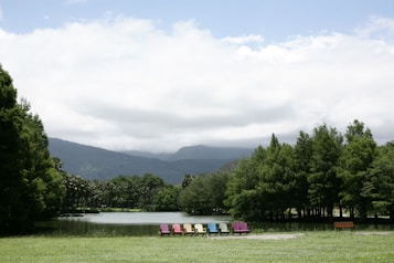 A serene landscape featuring a tranquil lake surrounded by lush green trees under a partly cloudy sky. A row of colorful Adirondack chairs is positioned on the grassy bank, adding vibrant contrast to the natural scenery. Distant mountains provide a majestic backdrop, enhancing the peacefulness of the setting.