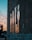 A series of pixelated figures are crafted using mirror tiles attached to a rough, textured wall. The figures reflect the sunset colors creating a warm glow. In the distance, blurred lights and the silhouette of a tree are visible, enhancing the ambient atmosphere.
