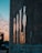 A series of pixelated figures are crafted using mirror tiles attached to a rough, textured wall. The figures reflect the sunset colors creating a warm glow. In the distance, blurred lights and the silhouette of a tree are visible, enhancing the ambient atmosphere.
