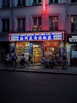 A street view of a Chinese restaurant with bright neon signs and outdoor seating. People are enjoying meals at wooden tables along the sidewalk during the evening. The building is lit up with colorful lights, creating a cozy atmosphere.