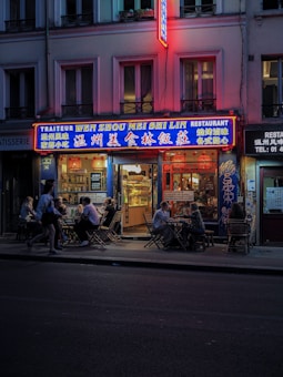 A street view of a Chinese restaurant with bright neon signs and outdoor seating. People are enjoying meals at wooden tables along the sidewalk during the evening. The building is lit up with colorful lights, creating a cozy atmosphere.