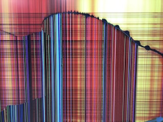 A screen display with multi-colored vertical and horizontal lines forming a pattern. The colors include reds, yellows, blues, and blacks. There are visible cracks along the surface, indicating a damaged or broken screen.