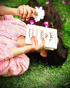 A person lies on the grass, holding a white flower and a book titled 'Girl in Pieces' by Kathleen Glasgow. The person has henna designs on their hands and wears a pink-patterned garment. Pink flower petals are scattered around.