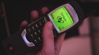 A hand holding a classic mobile phone with a green backlit screen displaying a menu. The phone has a physical keypad with numbers and icons, and the screen shows options like 'Services' and 'Home'. The device has a retro design with an antenna.