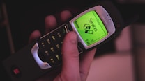 A hand holding a classic mobile phone with a green backlit screen displaying a menu. The phone has a physical keypad with numbers and icons, and the screen shows options like 'Services' and 'Home'. The device has a retro design with an antenna.
