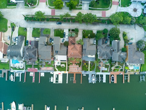 An aerial view of a row of houses along a waterfront, with neatly arranged docks and boats anchored. The neatly organized residential area shows various roofs, driveways, and green lawns. The image includes roads, parked cars, and landscaped gardens in front of the homes.