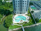 A high-rise building with a modern design is situated near a body of water. The front area features a turquoise swimming pool with a curved shape surrounded by neatly landscaped greenery and palm trees. Lounge chairs and outdoor furniture are arranged around the pool. The surrounding area includes tennis courts, additional buildings, and dense green trees.