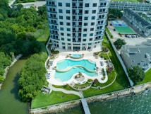 A high-rise building with a modern design is situated near a body of water. The front area features a turquoise swimming pool with a curved shape surrounded by neatly landscaped greenery and palm trees. Lounge chairs and outdoor furniture are arranged around the pool. The surrounding area includes tennis courts, additional buildings, and dense green trees.