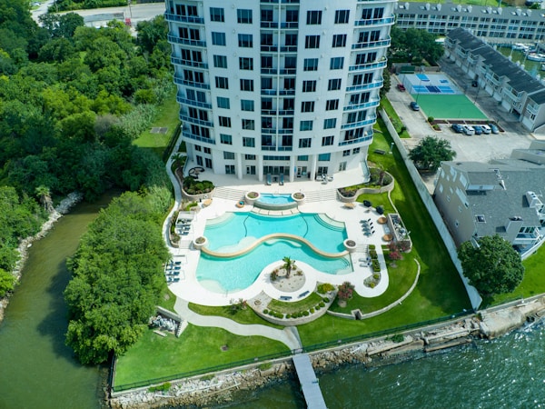 A high-rise building with a modern design is situated near a body of water. The front area features a turquoise swimming pool with a curved shape surrounded by neatly landscaped greenery and palm trees. Lounge chairs and outdoor furniture are arranged around the pool. The surrounding area includes tennis courts, additional buildings, and dense green trees.