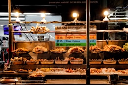 A street food stall displays a variety of skewered foods under bright lights. The skewers are carefully arranged on metal trays at different levels. A sign with pricing information and color-coded categories is prominently displayed behind the skewers. The stall has a bustling background, suggesting a vibrant market environment.