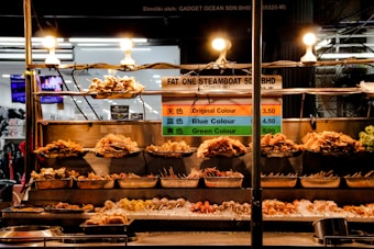 A street food stall displays a variety of skewered foods under bright lights. The skewers are carefully arranged on metal trays at different levels. A sign with pricing information and color-coded categories is prominently displayed behind the skewers. The stall has a bustling background, suggesting a vibrant market environment.