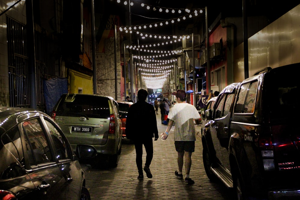 a couple of people walking down a street next to parked cars