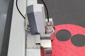 A machine or industrial device with metallic components, including a cylindrical part and wiring, is positioned near a pink material with circular cutouts on a gray surface.