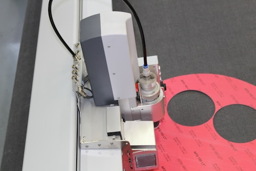 A machine or industrial device with metallic components, including a cylindrical part and wiring, is positioned near a pink material with circular cutouts on a gray surface.