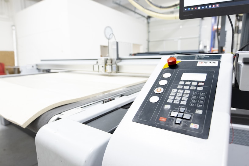 A close-up view of an industrial cutting machine in a manufacturing setting. There is a large control panel with numerical buttons, function keys, and a prominent red emergency stop button. The machine is processing a large sheet of material, likely for cutting or engraving. Overhead lighting and a computer monitor are visible in the background.