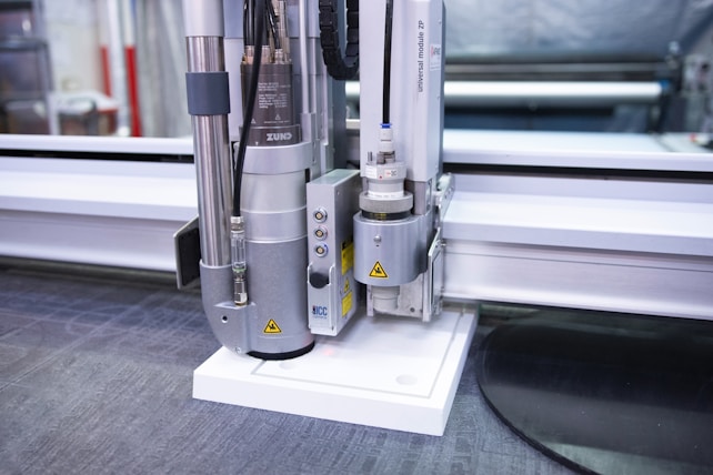 A close-up view of an industrial cutting machine with a metallic arm and attachments, positioned above a flat surface. The setting appears to be a workshop or manufacturing environment with additional machinery in the background.