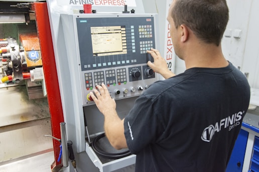 A person operates a CNC machine, focusing on the control panel with numerous buttons and a screen displaying data. The individual's hands are interacting with the controls, suggesting an adjustment or programming task. The surrounding environment appears to be a workshop or manufacturing setting.