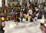 A festive scene featuring miniature holiday village decorations with various figures and buildings. In the center, a snowman in a purple vest and yellow boots appears to interact with two small figures. Snow-covered trees and vibrant lights add to the cheery ambiance, with a sign for 'Santa Claus & Pals' and 'Deb's Puppy Shelter' visible in the background. People are engaged in various activities, creating a lively winter village atmosphere.