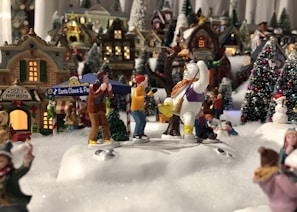 A festive scene featuring miniature holiday village decorations with various figures and buildings. In the center, a snowman in a purple vest and yellow boots appears to interact with two small figures. Snow-covered trees and vibrant lights add to the cheery ambiance, with a sign for 'Santa Claus & Pals' and 'Deb's Puppy Shelter' visible in the background. People are engaged in various activities, creating a lively winter village atmosphere.