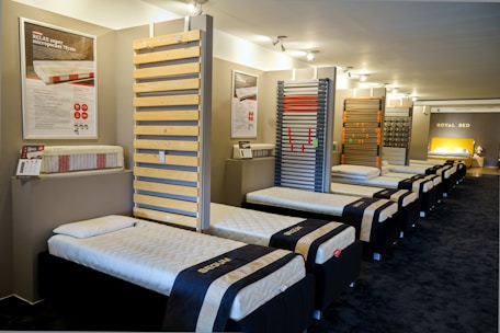 The interior of a showroom displaying several neatly arranged single beds lined up in a row. The beds have white mattresses and dark frames. Informative posters and samples are mounted on the walls, and there is a bright focus on a featured section at the end of the room labeled 'Royal Bed.'