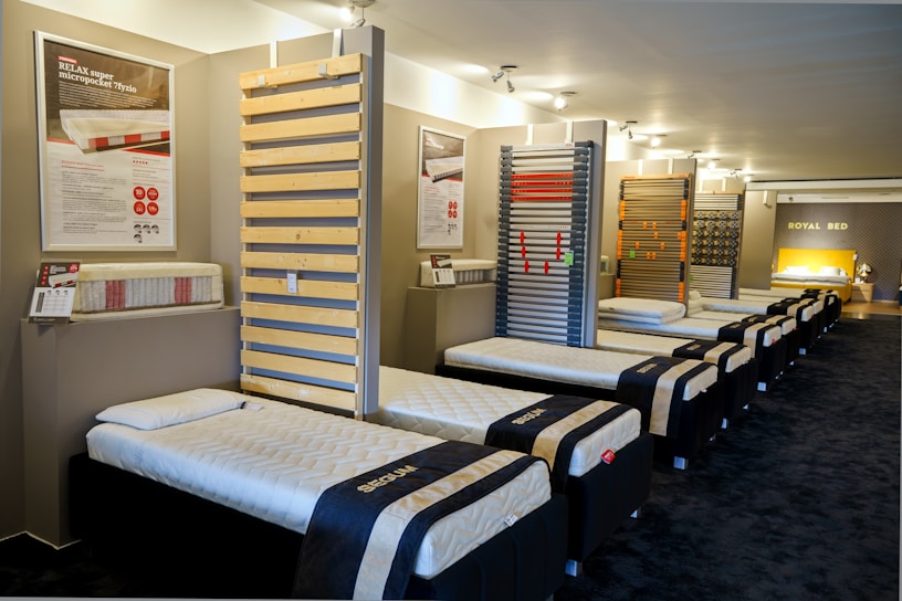 The interior of a showroom displaying several neatly arranged single beds lined up in a row. The beds have white mattresses and dark frames. Informative posters and samples are mounted on the walls, and there is a bright focus on a featured section at the end of the room labeled 'Royal Bed.'