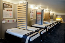 The interior of a showroom displaying several neatly arranged single beds lined up in a row. The beds have white mattresses and dark frames. Informative posters and samples are mounted on the walls, and there is a bright focus on a featured section at the end of the room labeled 'Royal Bed.'