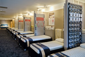 A showroom with multiple rows of neatly arranged bed frames and mattresses. The beds are all aligned in a clean, spacious environment with a modern design. The walls display various bed-related equipment and informational posters.