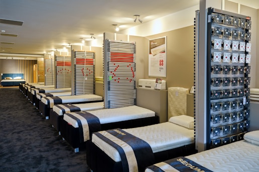 A showroom with multiple rows of neatly arranged bed frames and mattresses. The beds are all aligned in a clean, spacious environment with a modern design. The walls display various bed-related equipment and informational posters.