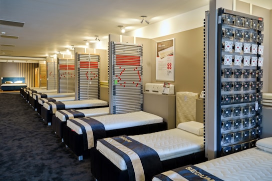 A showroom with multiple rows of neatly arranged bed frames and mattresses. The beds are all aligned in a clean, spacious environment with a modern design. The walls display various bed-related equipment and informational posters.