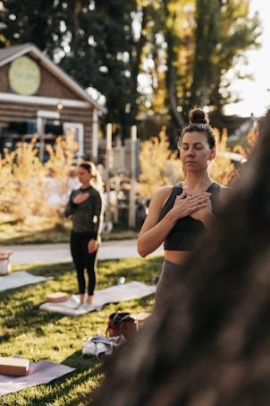 Two individuals are practicing a yoga or meditation session outdoors. The person in the foreground is standing with eyes closed, hands over the heart, conveying a sense of calm and focus. The background features a wooden building and autumn trees with golden leaves, under a soft, warm light.