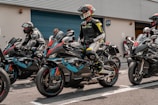 A group of motorcyclists in full leather gear are seated on racing motorcycles, preparing for a race. The riders are wearing helmets with visors down, and their bikes are adorned with sporty designs and brand logos. They are positioned in what appears to be a pit lane next to a garage or storage unit. The scene conveys a sense of anticipation and readiness for action.