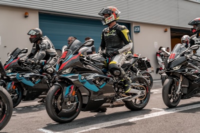 A group of motorcyclists in full leather gear are seated on racing motorcycles, preparing for a race. The riders are wearing helmets with visors down, and their bikes are adorned with sporty designs and brand logos. They are positioned in what appears to be a pit lane next to a garage or storage unit. The scene conveys a sense of anticipation and readiness for action.