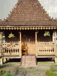 A traditional wooden structure with an intricately designed roof, featuring geometric patterns along the edges. There are two hanging potted plants with greenery on the veranda, and a wooden chair placed in front of the door. The structure is elevated with steps leading up to the entryway.