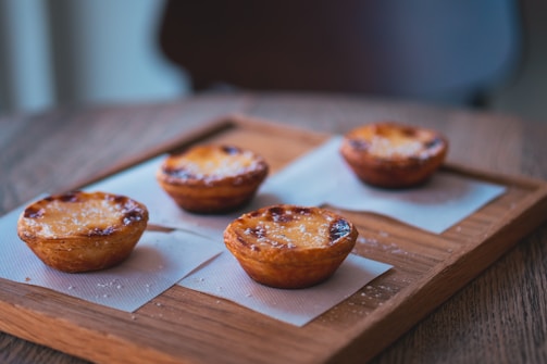 Four golden-brown pastries are neatly arranged on a wooden tray, each placed on a square napkin. The pastries have a slightly caramelized crust with scattered sugar crystals visible on their tops. The setting conveys a warm, cozy atmosphere with natural lighting gently highlighting the textures of the food and tray.