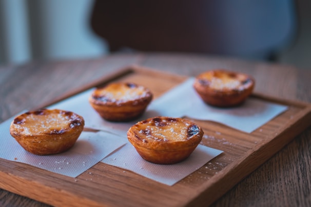 Four golden-brown pastries are neatly arranged on a wooden tray, each placed on a square napkin. The pastries have a slightly caramelized crust with scattered sugar crystals visible on their tops. The setting conveys a warm, cozy atmosphere with natural lighting gently highlighting the textures of the food and tray.