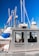 A metal emergency boat equipped with antennas and communication devices docked at a marina with several sailboats in the background. The boat has windows and is labeled with 'Emergencies Urgences' on its side. A flag is attached to one of the boat's antennas.
