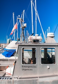 A metal emergency boat equipped with antennas and communication devices docked at a marina with several sailboats in the background. The boat has windows and is labeled with 'Emergencies Urgences' on its side. A flag is attached to one of the boat's antennas.