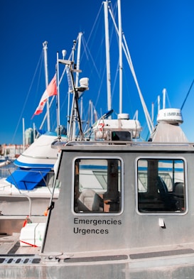 A metal emergency boat equipped with antennas and communication devices docked at a marina with several sailboats in the background. The boat has windows and is labeled with 'Emergencies Urgences' on its side. A flag is attached to one of the boat's antennas.