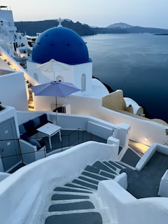 A scenic view of a coastal area featuring whitewashed buildings with blue domes, characteristic of Cycladic architecture. A staircase leads down to a seating area with a table and chairs positioned under a blue umbrella. The tranquil sea stretches out towards the horizon with rugged hills in the distance.