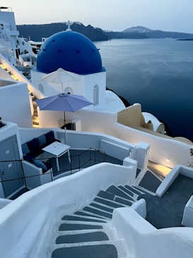 A scenic view of a coastal area featuring whitewashed buildings with blue domes, characteristic of Cycladic architecture. A staircase leads down to a seating area with a table and chairs positioned under a blue umbrella. The tranquil sea stretches out towards the horizon with rugged hills in the distance.