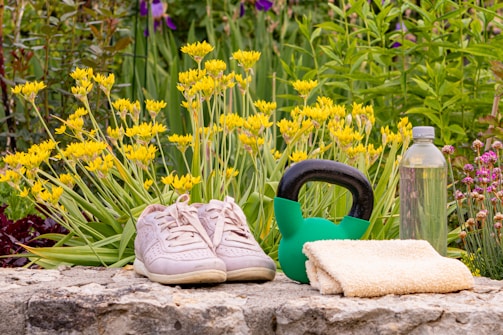Two pink sneakers, a green kettlebell, a small towel, and a clear water bottle are arranged on a stone surface. Behind them, yellow flowers and green foliage create a vibrant natural background.