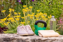 Two pink sneakers, a green kettlebell, a small towel, and a clear water bottle are arranged on a stone surface. Behind them, yellow flowers and green foliage create a vibrant natural background.