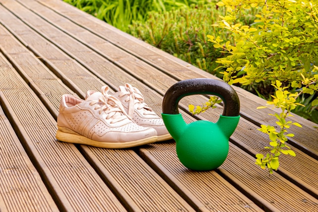 A pair of light-colored athletic shoes are placed on a wooden deck beside a green kettlebell. The background includes vibrant green foliage, adding a fresh outdoor atmosphere.