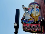 A vintage-style neon sign featuring a cowgirl playing an electric guitar is mounted on a brick wall. The sign is lit up with various colors, and next to it stands a tall streetlamp with a decorative top against a clear blue sky.