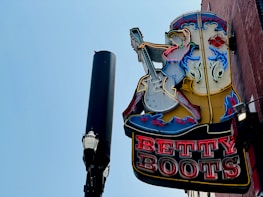 A vintage-style neon sign featuring a cowgirl playing an electric guitar is mounted on a brick wall. The sign is lit up with various colors, and next to it stands a tall streetlamp with a decorative top against a clear blue sky.