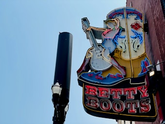 A vintage-style neon sign featuring a cowgirl playing an electric guitar is mounted on a brick wall. The sign is lit up with various colors, and next to it stands a tall streetlamp with a decorative top against a clear blue sky.