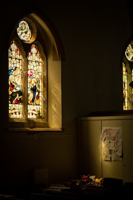 A dimly lit room with tall stained glass windows featuring intricate designs and figures casts colorful light within. Below the window, there is a cluttered area displaying various toys and drawings on a cabinet, indicating a space used for activities or gatherings.