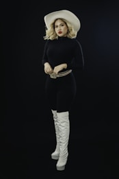 A person wearing a white cowboy hat, black outfit, and high white boots stands confidently. Their hands rest on a decorative belt buckle, and they have red lipstick and wavy blonde hair.